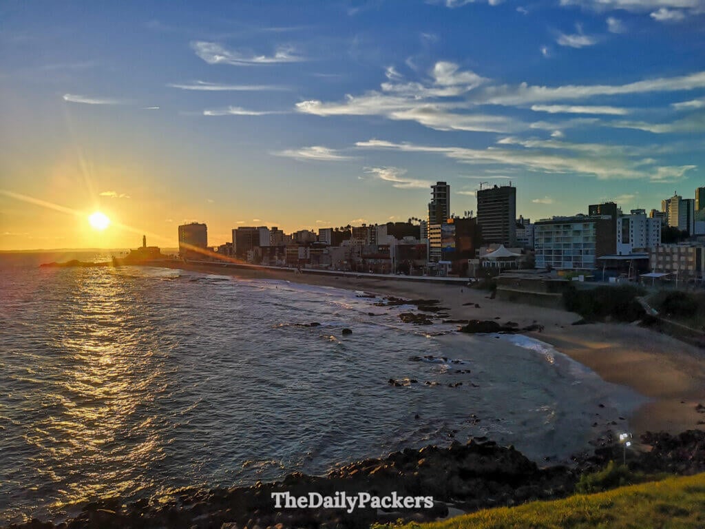 Sunset view from Morro do Cristo in Salvador with the Atlantic Ocean, rocky coastline, city skyline, and the lighthouse glowing at golden hour