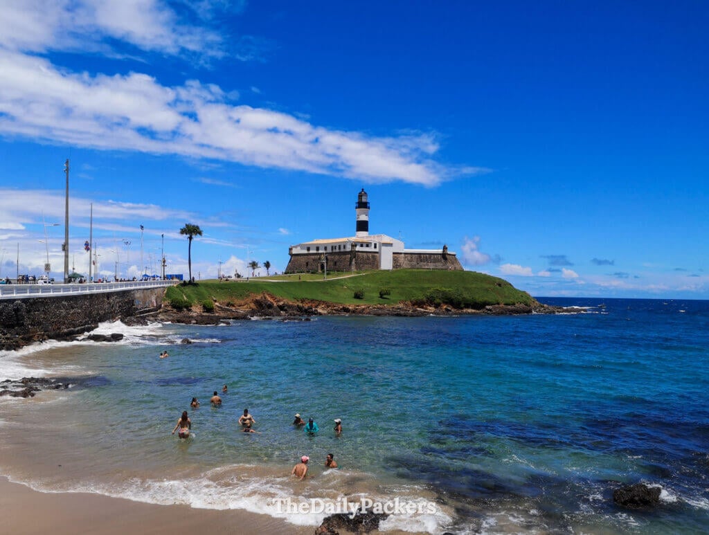 People swimming at Farol da Barra beach in Salvador with the lighthouse and fort rising above the rocky shoreline