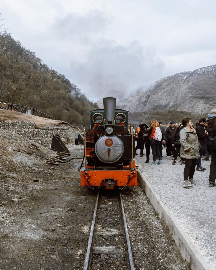 Historic steam train in Ushuaia, Argentina with a crowd of tourists and scenic valley views.
