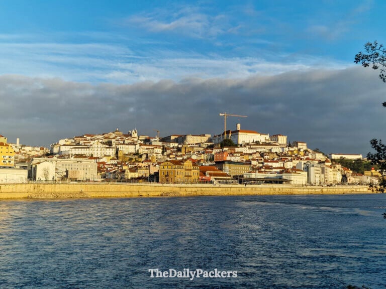 Panoramic view of Coimbra's historic hillside and the Mondego River under a blue sky at sunset.