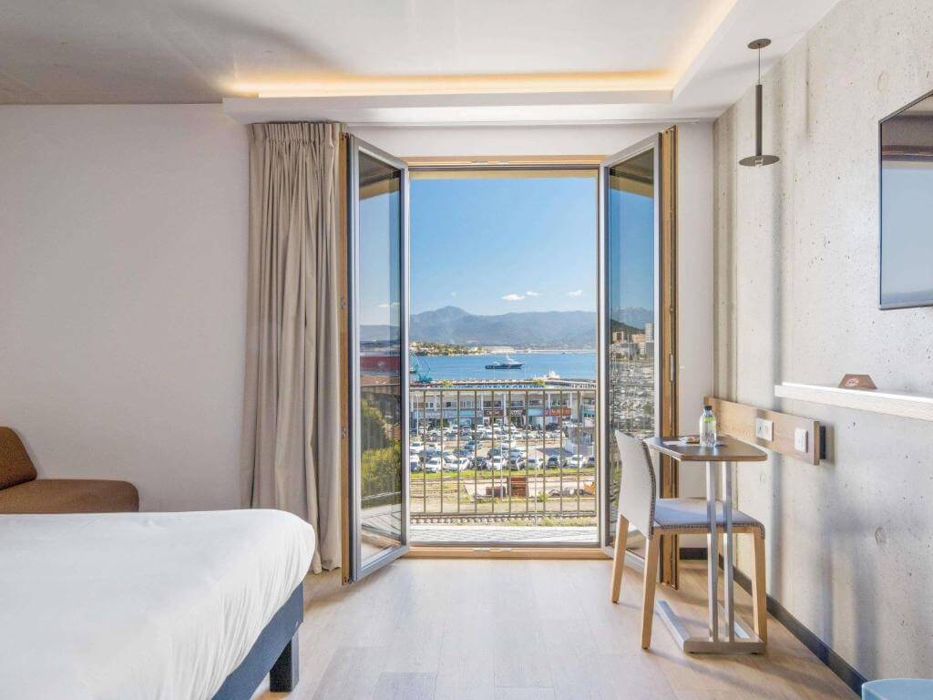 Modern room at Ibis Styles Ajaccio Napoleon featuring a balcony and a panoramic view of Ajaccio city and the harbor.
