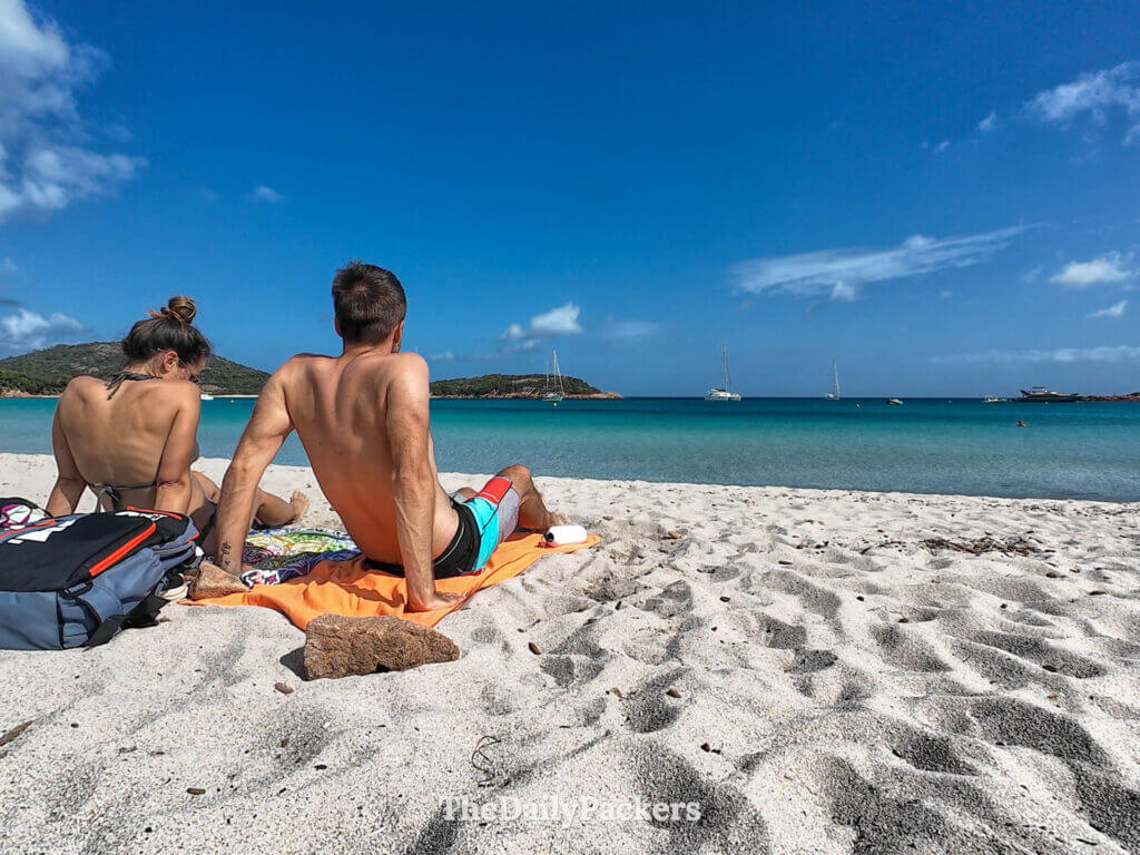 A couple sitting together on the white sand looking at the turquoise water of Rondinara Beach.
