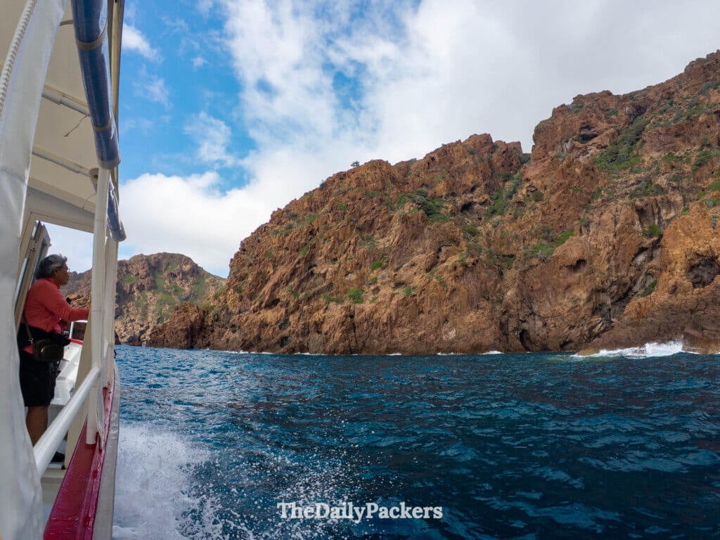 Tour boat navigating past massive orange volcanic rock cliffs in Scandola Reserve, Corsica.
