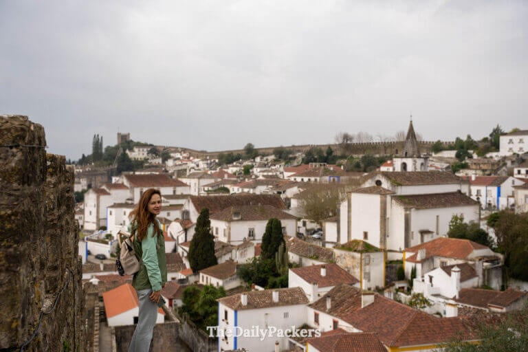 A woman poses on the medieval ramparts with the white village and Obidos Castle behind her.