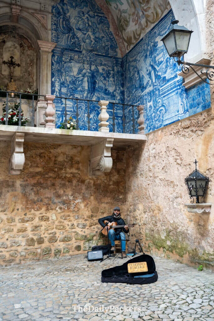 A street musician plays guitar under the blue azulejo tiled arches of Porta da Vila.