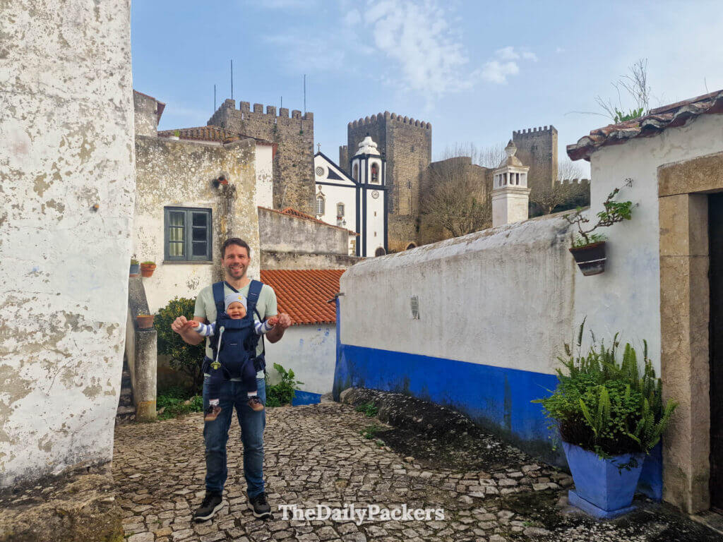 A father carrying his baby in a harness down a scenic backstreet near Óbidos Castle.