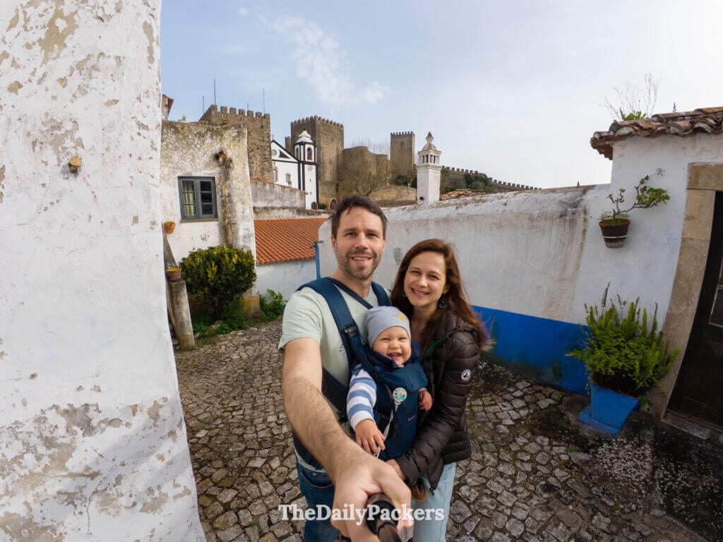 Happy family selfie on a historic cobblestone street with Óbidos Castle behind them.