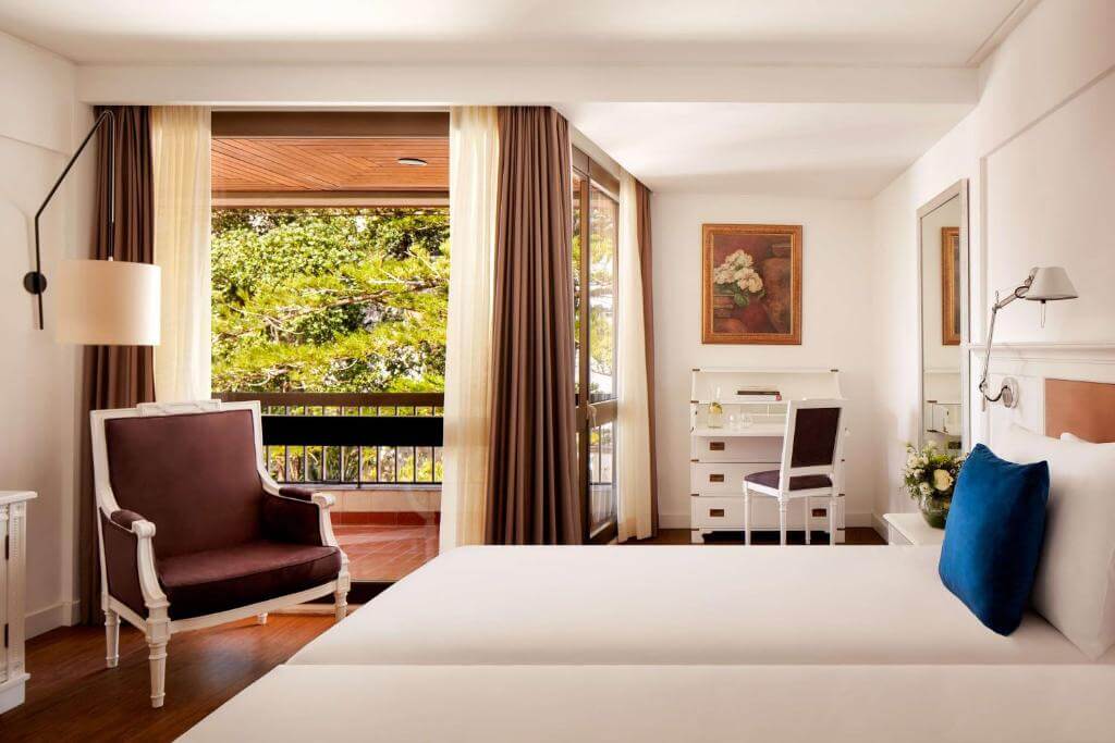 Elegant hotel room with an armchair, desk, and balcony overlooking green trees.