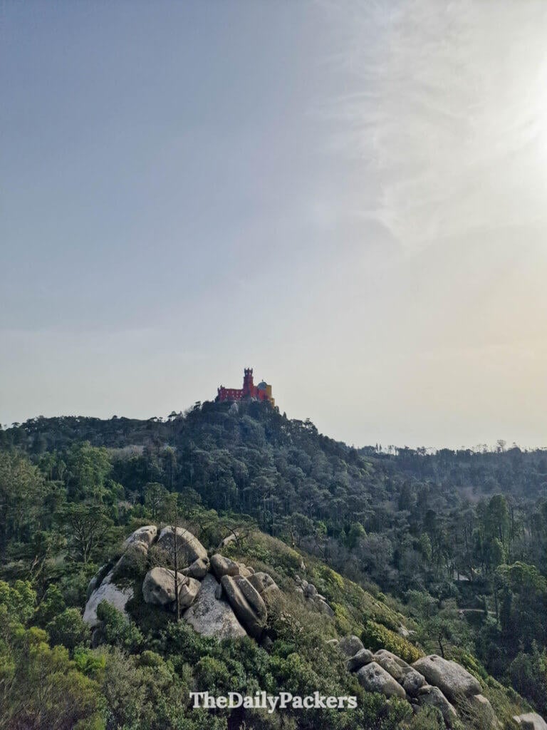 Distant view of the colorful Pena Palace standing atop a lush, forested hill in Sintra.