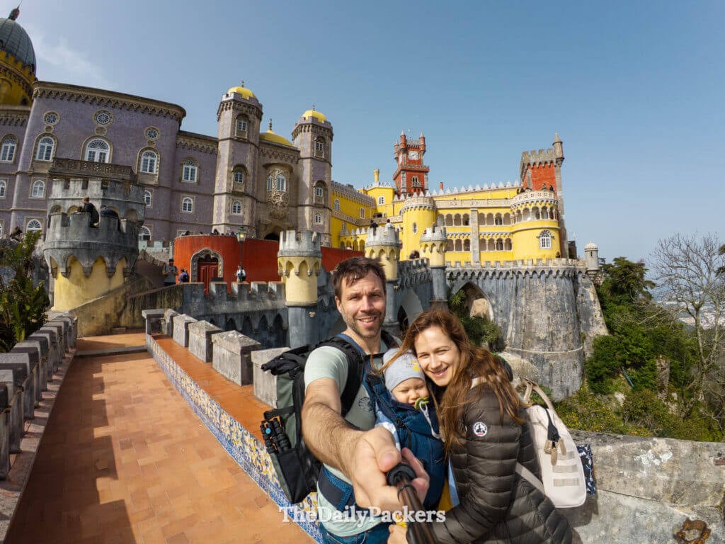 Family selfie with a baby on a stone walkway overlooking the colorful facades of Pena Palace.